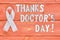White awareness ribbon and inscription of iron letters: thanks doctors day lying on wooden textured background color of season