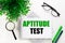 On a white background lies a notebook with the word APTITUDE TEST, glasses, a magnifying glass, green markers and a green plant