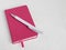 White ballpoint pen on a closed notebook in a pink cover on a white background