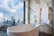 White bathtub near the window, overlooking the cityscape, building landscape outside scenery. Luxury master bathroom interior
