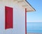 White beach hut with red shutter, blue sea and sky.