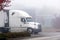 White big rig semi truck with refrigerated semi trailer leaves the warehouse after loading cargo going on foggy road