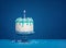 White birthday drip cake with teal ganache and lit candle over dark blue background