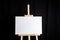 White blank canvas stands on a wooden artistic easel on black curtain background. Horizontal rectangular mockup canvas wrapped