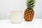 A white blank enamel mug standing out on a white table with a whole pineapple close it it