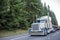 White and blue big rig classic American semi truck with flat bed semi trailer driving on the road with frosted winter evergreen