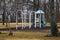 A white and blue jungle gym and a swing set in the park surrounded by yellow winter grass and bare winter trees