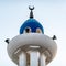 White and blue painted tip of a minaret with loudspeakers and a bronze crescent moon on the side.