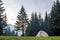 White and blue tourist tent on green meadow between evergreen fir-trees forest with beautiful mountain in distance. Tourism, outdo