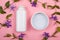White bottles with body cosmetics on a pink background with branches of purple wildflowers top view. skin care, beauty