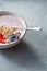 White bowl with homemade granola, sliced strawberries, yogurt, blueberry, spoon - natural breakfast on a gray table.