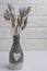 White brick background with a light two-color vase with a plaster heart on the neck and dried hare tails. Scandinavian interior de