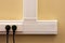 White cable channel with electrical outlets on a yellow wall, where two black plugs are inserted.