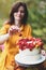 White cake with berries, maple leaves, apples, rowan. Woman in yellow dress on wooden chair by round table with blue tablecloth,