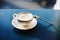 A white cappuccino Cup on a saucer with a teaspoon on a blue table