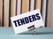 A white card with the text TENDERS stands on a clip for papers on the table against the background of books. Defocus