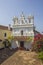 White catholic church next to the yellow house and tropical plants under the blue sky