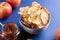 White ceramic cup with apple chips, fresh red apples and glass jar of apple jam on blue background