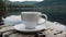 A white coffee cup on a saucer sits on a wooden surface near a lake