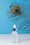 A white cosmetic matte jar with a pipette stands on a blue background with a peacock feather. Vertical photo. Container for oil, s