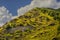 White cross mounted half way up a mountain where its eroding with trees farther up under blue cloudy sky