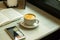 White Cup with Cappuccino on Table with Open Book Smartphone on Cafe Table by Window.