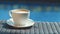 White cup of cappuccino on wicker table with blue water background