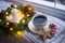 White cup of coffee and Christmas gingerbread near fir wreath decorated with red balls, burning candle and coiled with glowing gar