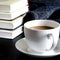 A white cup with a fragrant cove stands on a black table next to books and a blue wool scarf