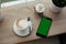 White cup of hot cappuccino on white saucer, black mobile phone, red velvet dessert and green grass in pots on wooden bar table