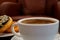 White cup of hot coffee with light smoke, blurred doughnuts and armchair in background