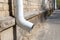 White downpipe on the wall of a brick building in dry, sunny weather. Tube, gutters. Drainpipe and asphalt sidewalk