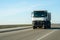 A white dump truck drives out of town on a flat asphalt road among fields against a blue sky on a sunny day