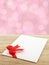 white envelope of greeting card with simple red ribbon bow on wooden table top with defocused pastel pink lights bokeh