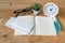 White hygienic cloth mask on opened notebook, alcohol in spray bottle, glasses , and plastic green tree on wooden desk