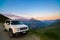 White Jeep Renegade parked on dirt road at panoramic view point on the Italian Alps from above. Colorful sky at sunset, mist on th