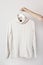 White knitted wool sweater on a white wooden hanger in female hands against the background of the light wall. Autumn and winter cl