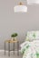 White lamp above leaf in vase on wooden bedside table in scandinavian bedroom, copy space on the empty grey wall