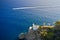 White lighthouse is on the top of the hill in Ligurian Sea with the sailing white motor boat on the background. Arial view on the