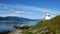 White lighthouse tower with red top and moss covered rocky shore of a Norwegian Fjord on a sunny day, with mountains, blue sky and