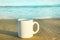 White Mock Up Mug with Empty Space for Artwork Text Standing on Beach Sand. Turquoise Blue Sea