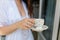 White mug with a pattern with hot coffee inside the in the hands of women in Bathrobe standing on the balcony of her bedroom. Woma