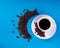 A white mug on a saucer with a black drink is set on a blue background with a number of scattered crescent coffee grains