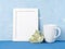 White mug with tea or coffee, frame, flower bouquet on blue table opposite blue concrete wall. Mock up with empty space