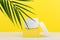 White opened cosmetic jar on yellow podium with palm leaf against bright yellow background.