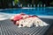 A white and orange Turkish towel, bikini top, and white seashells on rattan lounger with blue a swimming pool as background.