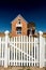 White picket fence gate boarders the land before a red brick house with dark sky