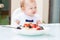 White plate bowl with oatmeal with blackberries and strawberries on a white table. Side view. In the background, a blurry baby is