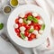 White plate of classic delicious caprese salad with ripe tomatoes, mozzarella and fresh basil leaves on gray concrete background.