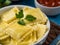 White plate with ravioli. A sprig of green mint lies on top. In the background is a photo of a bowl of sauce. Brown napkin. Blue